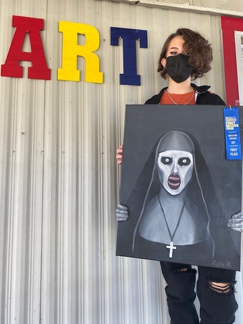 Zyla Bowman (6th) holds up her painting with a 1st place blue ribbon in front of a sign that says "ART"