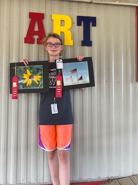 Abigail Moss (6th) holds up her photos with two 2nd place ribbons in front of a sign that says "ART"