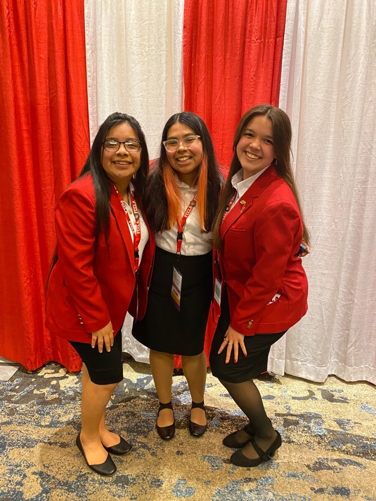 Pictured from left to right: Sarai Velasquez, Cinthia Rios, and Emma Derise are standing in front of a white and red striped back ground and wearing FCCLA official dress (white collared shirt with black pencil skirt and red blazer)